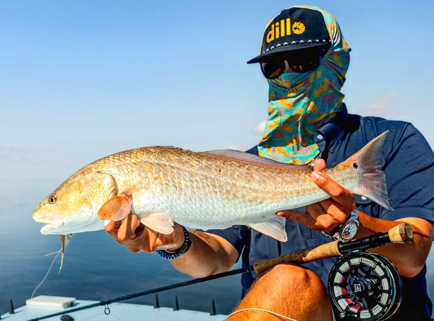 A fly fisherman wearing a colorful patterned neck gaiter, sunglasses, and a black Dill brand cap holds a large redfish on a boat under a clear blue sky.