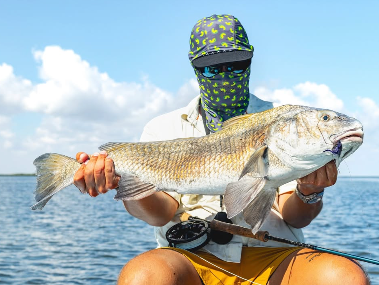 A person in a boat on the water wearing a green and yellow patterned face covering, sunglasses, and a white shirt holds a large redfish. A fly fishing rod is visible in their lap, with blue sky and water in the background.