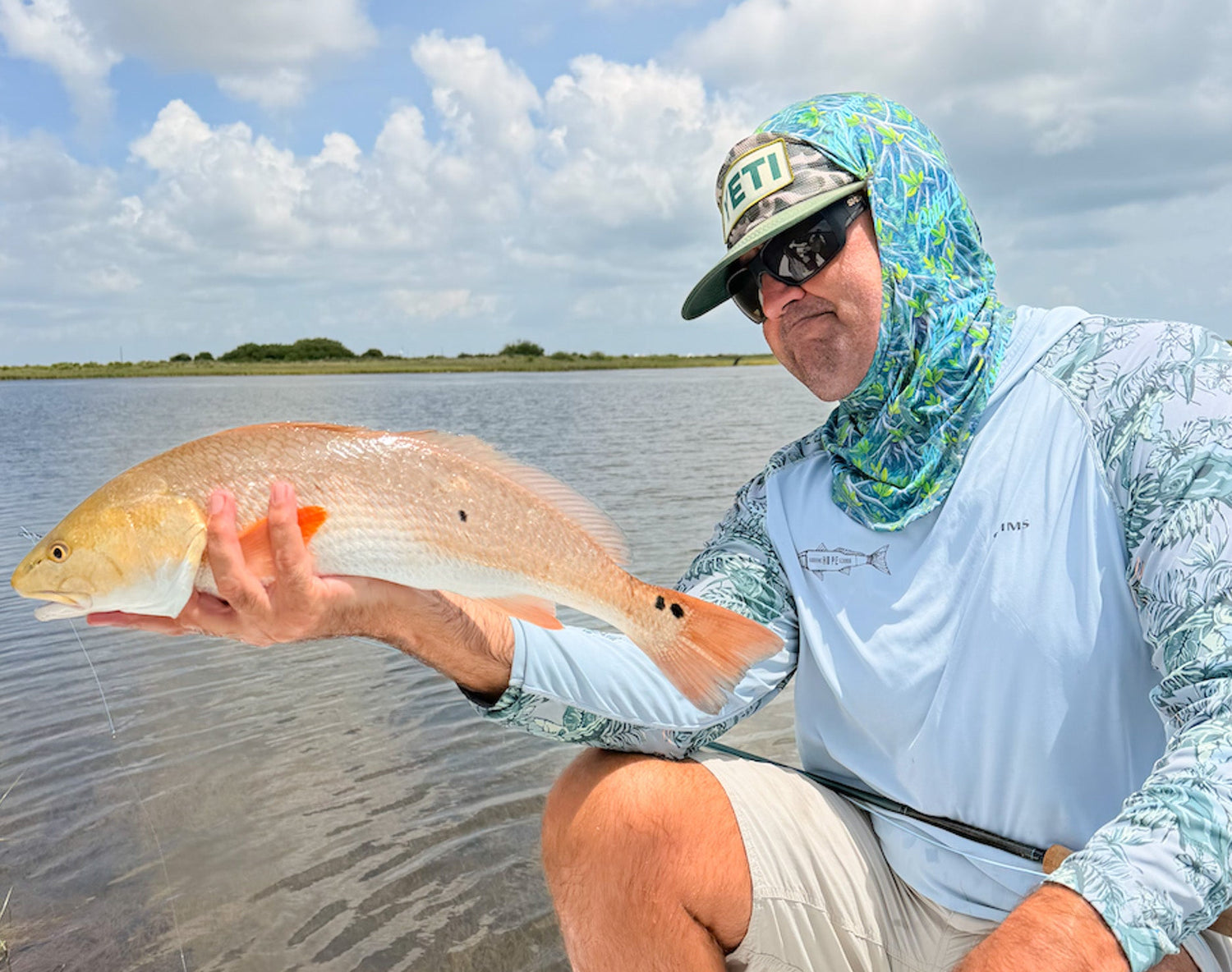 A man wearing a light blue long-sleeve fishing shirt, a green and blue patterned neck gaiter, and a cap sits in a boat holding a large redfish over water under a cloudy sky.