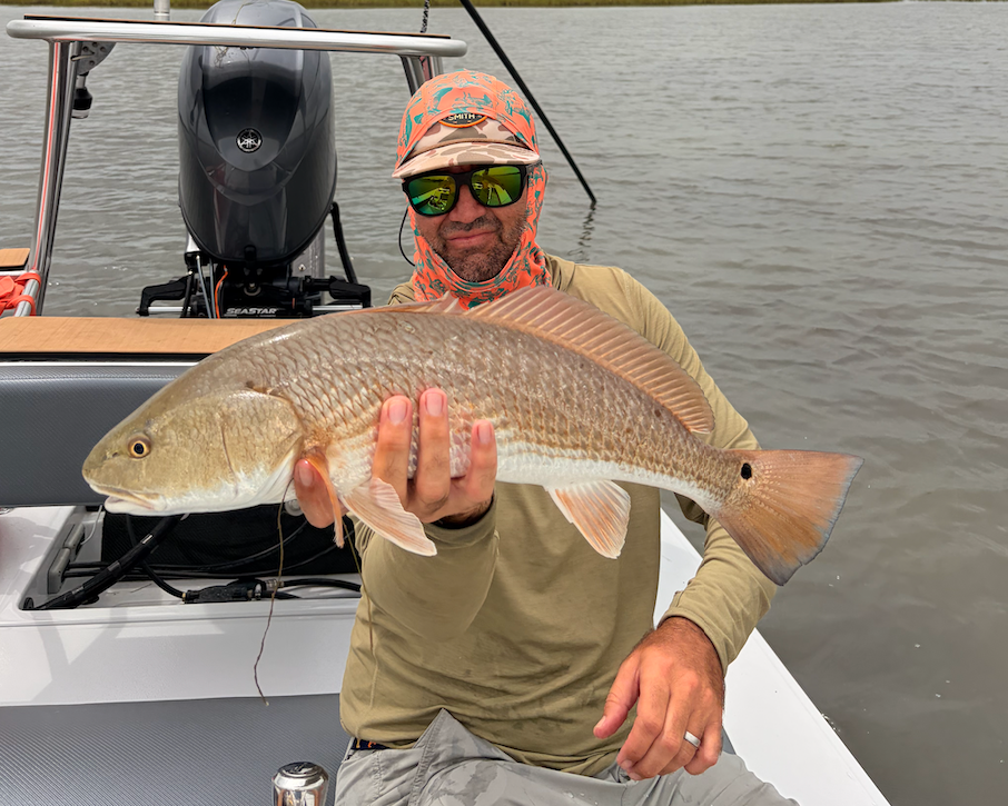 A man wearing a camouflage cap, orange patterned neck gaiter, and olive green long-sleeve shirt sits on a boat holding a large redfish. The background shows calm water, distant marshland, and an outboard motor under an overcast sky.